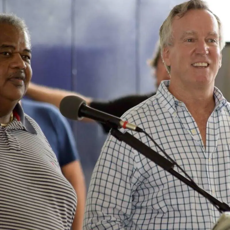 Two smiling men standing at a microphone during an outdoor gathering.