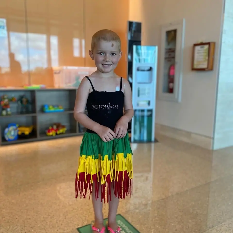 Smiling young child in a Jamaica-themed costume standing in a hospital lobby.
