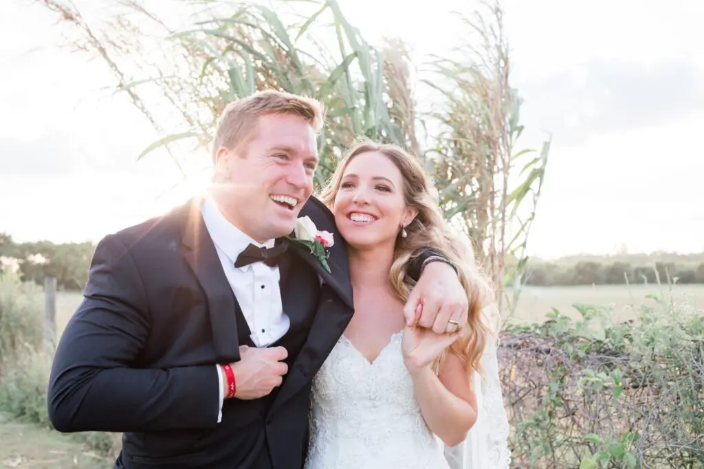 Smiling groom in tuxedo and bride in lace gown embrace outdoors at golden hour.