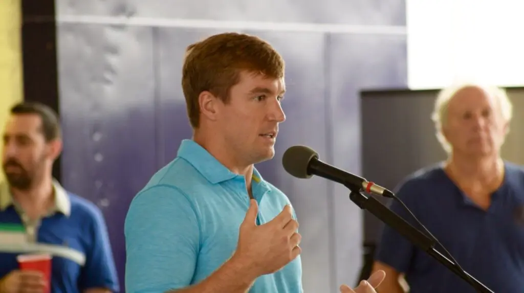 Red-haired man in a blue polo speaking into a microphone at an indoor event.