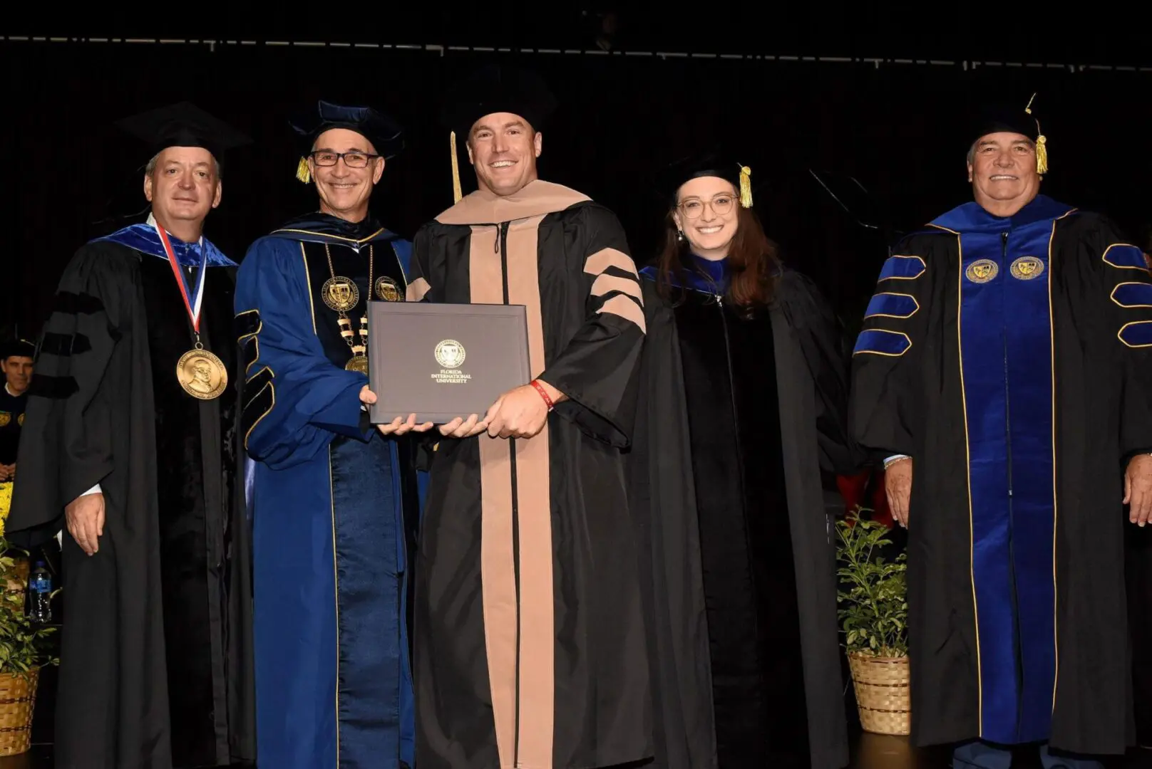 Five people in academic regalia present a doctoral diploma at an FIU commencement.