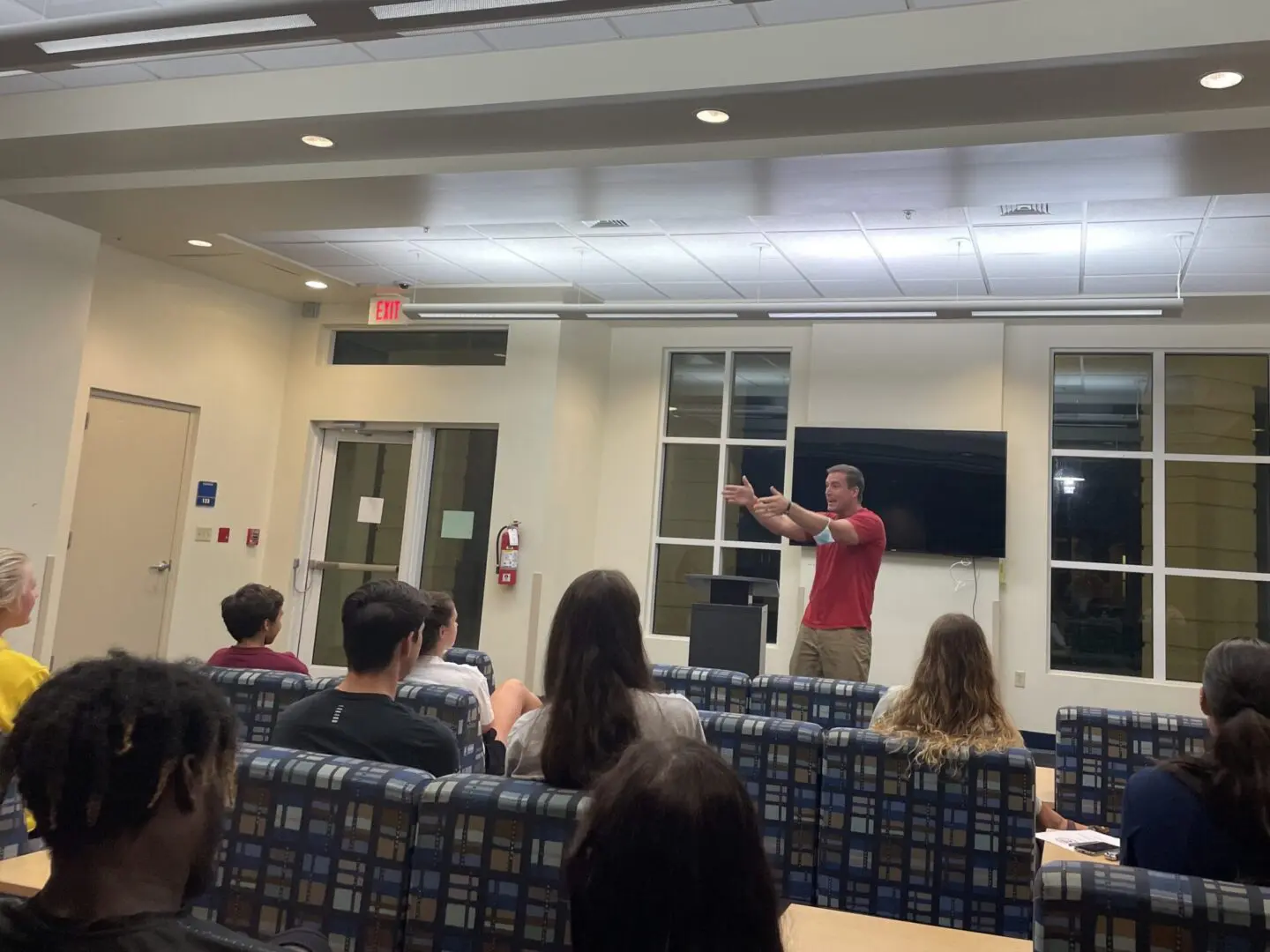 Man in a red shirt gestures enthusiastically while presenting to seated students in a classroom.