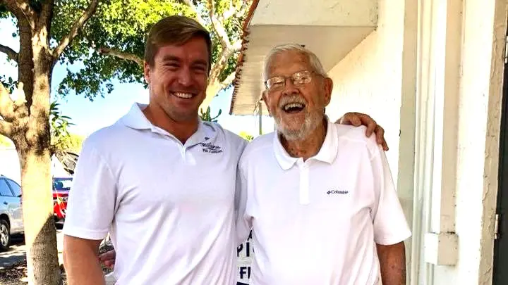 Two smiling men in white polos pose together outdoors, one young and one elderly.