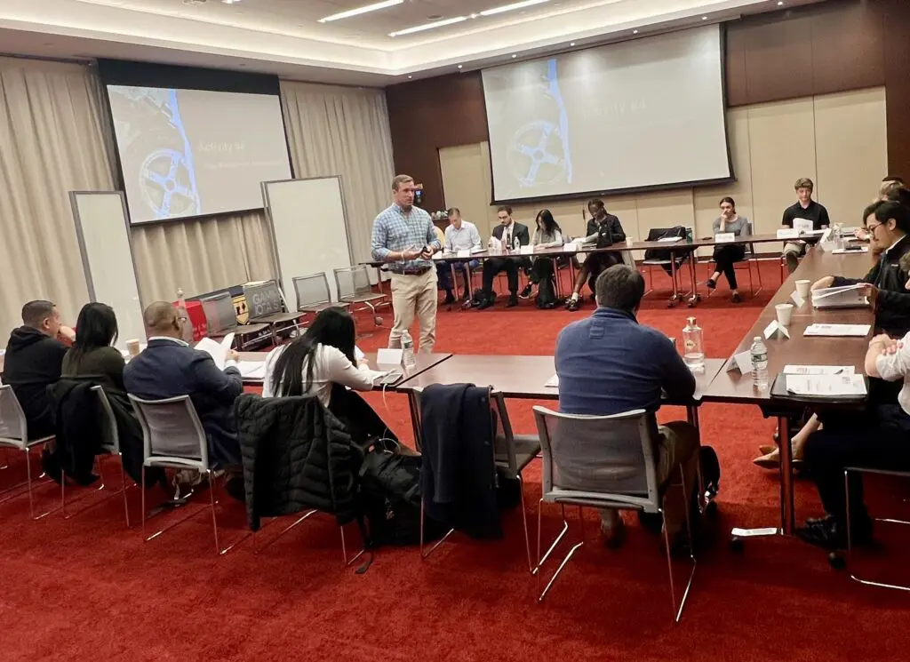 Speaker addressing participants seated around a U-shaped table in a workshop room.