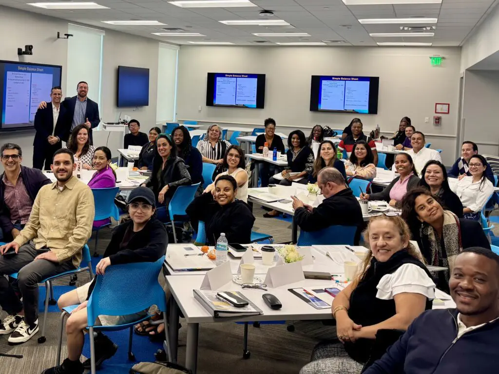 Diverse group of adult students smiling in a classroom with balance-sheet slides on screens.