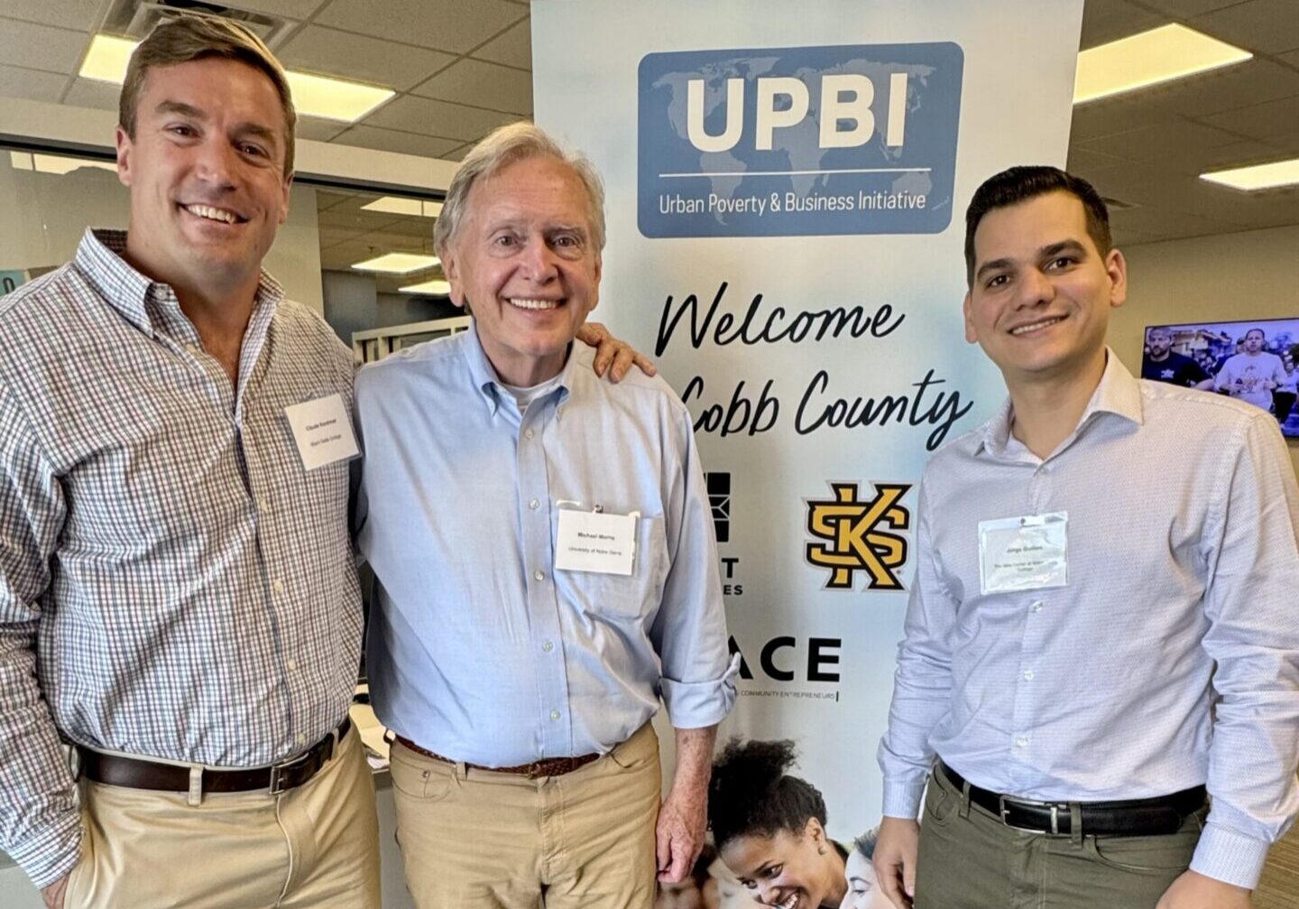 Three men smiling together in front of a UPBI Urban Poverty and Business Initiative banner.