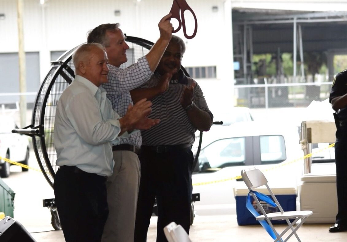Three men in business casual applaud during a ribbon-cutting ceremony.