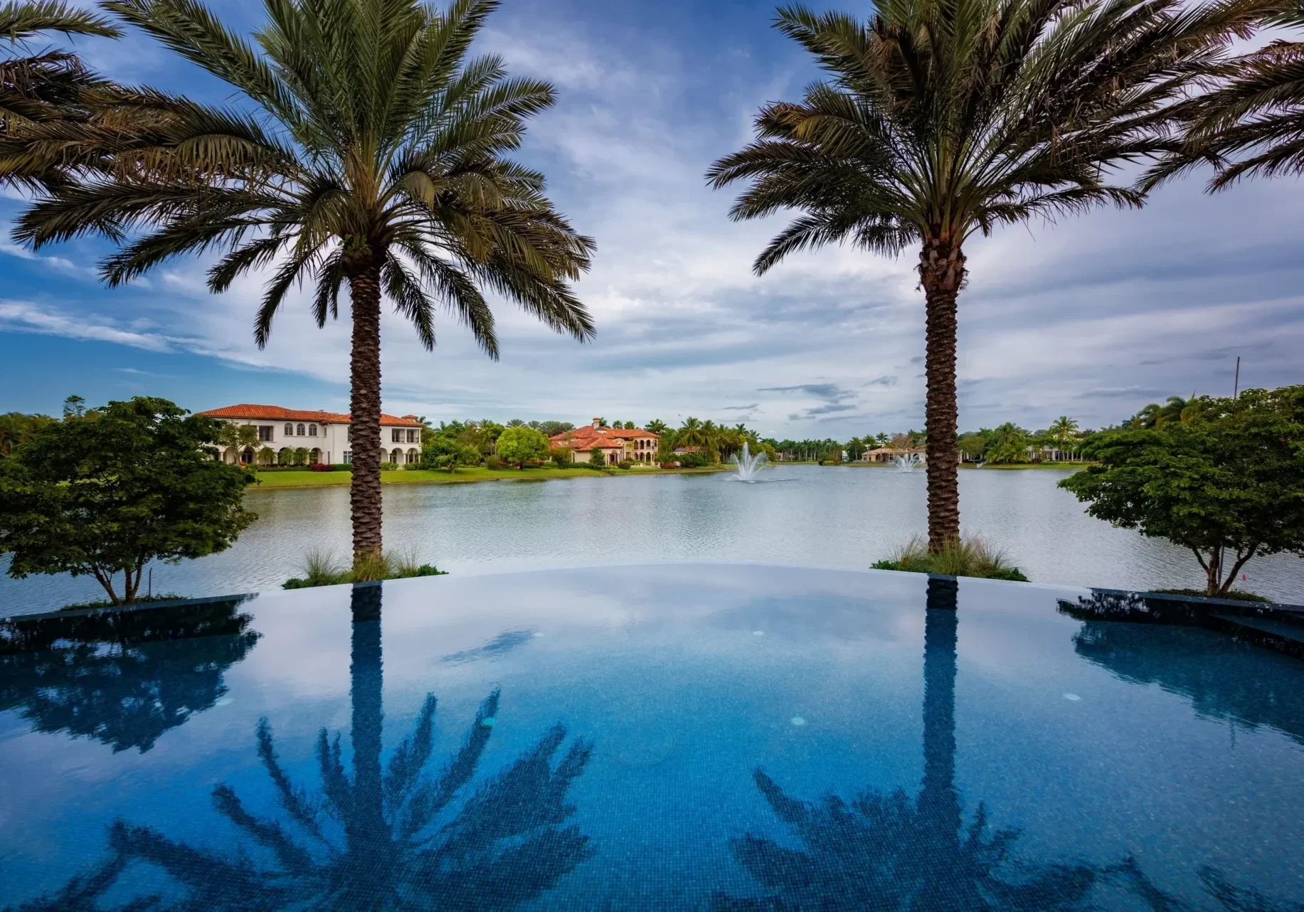 Infinity pool overlooking a lake with fountains and Mediterranean-style mansions framed by palms.
